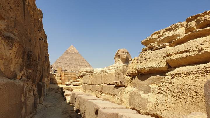 Iconic view of the Sphinx framed by sandstone walls with the Pyramid of Khafre rising in the background under a blue sky.