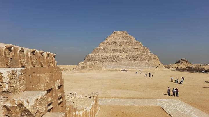 Wide open courtyard view of the Step Pyramid of Djoser with small groups of tourists scattered across the sand.