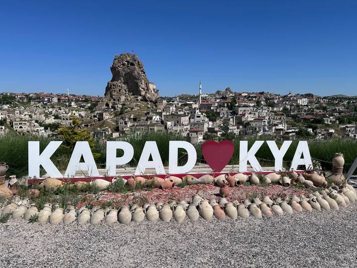 Large 'KAPADOKYA' sign with heart symbol frames Uchisar Castle and the town behind it under clear blue sky.