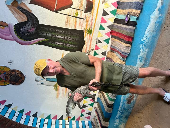 Man holding a baby crocodile inside a colorful Nubian-style room with painted murals