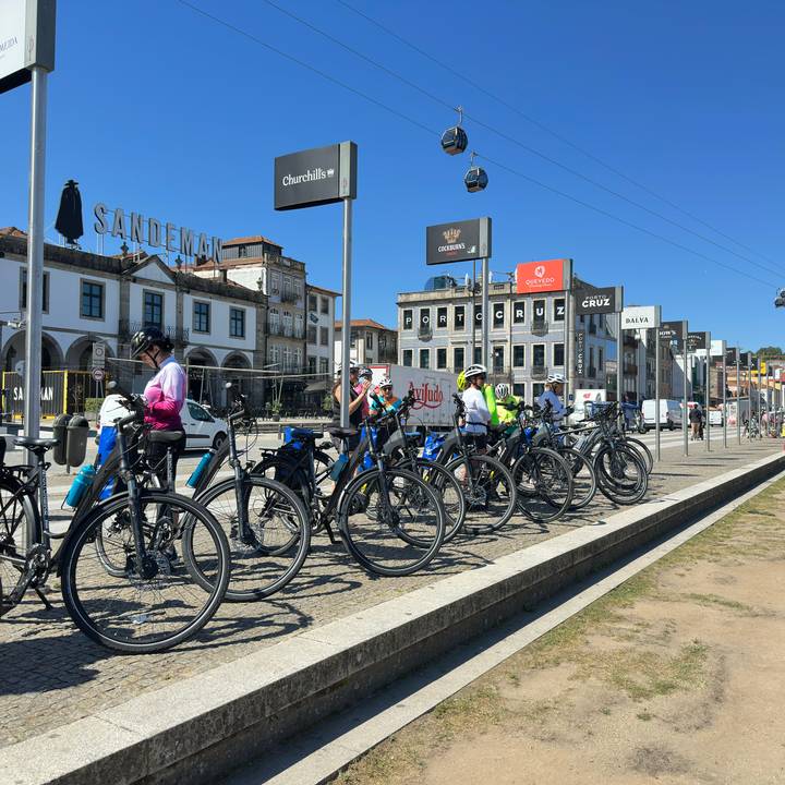Cyclists line up with their bikes near riverfront wine warehouses and signage in Porto.
