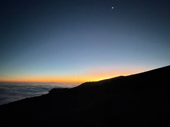 Silhouetted mountain ridge above a sea of clouds with a colorful sunrise glow on the horizon.