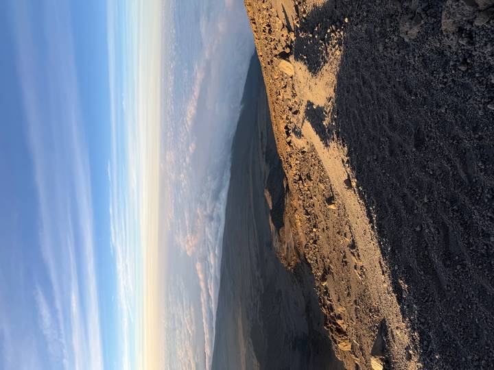 Rocky volcanic slope leading down to clouds with vast horizon and blue sky.