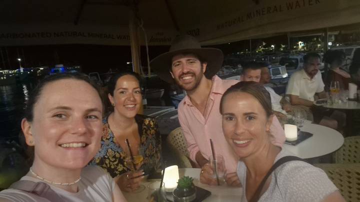 Group of friends take a low-light selfie at an outdoor waterfront bar in the evening.