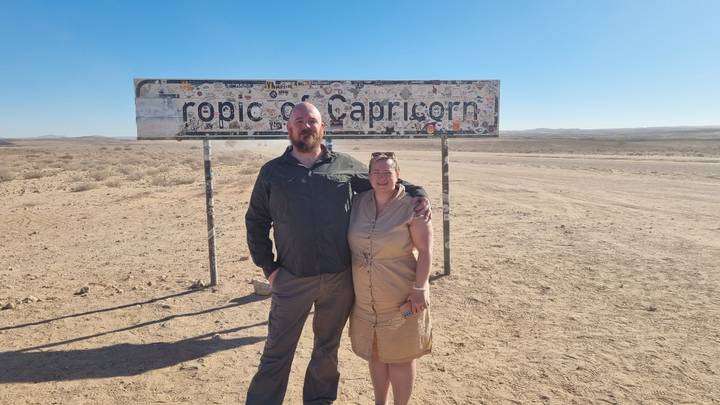 Couple standing by the sticker-covered Tropic of Capricorn sign on a wide, dusty desert plain under a cloudless blue sky.