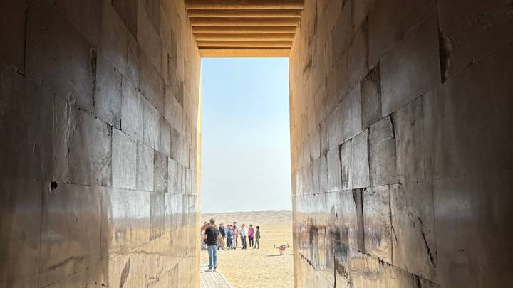 Long narrow stone corridor opening onto a bright desert plain where a tour group gathers at the exit.