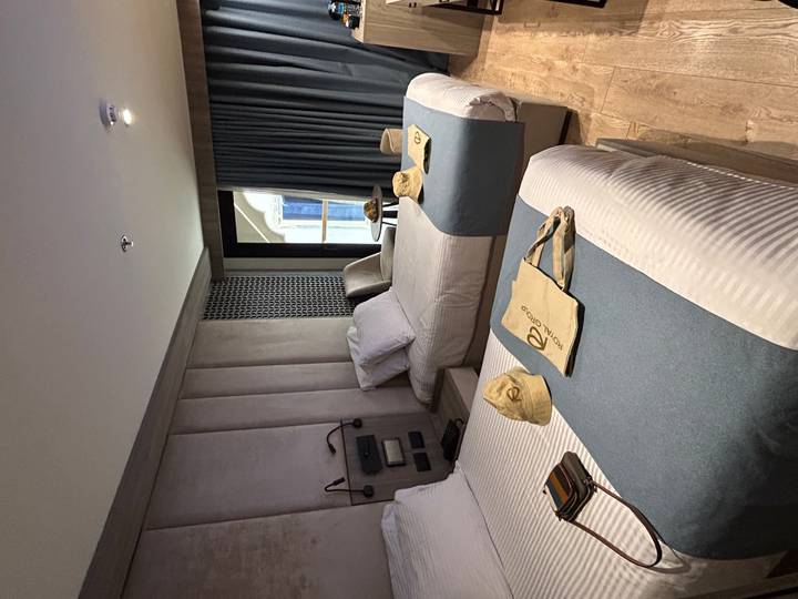 A neatly arranged cruise-ship cabin with two twin beds, teal throws and welcome bags.