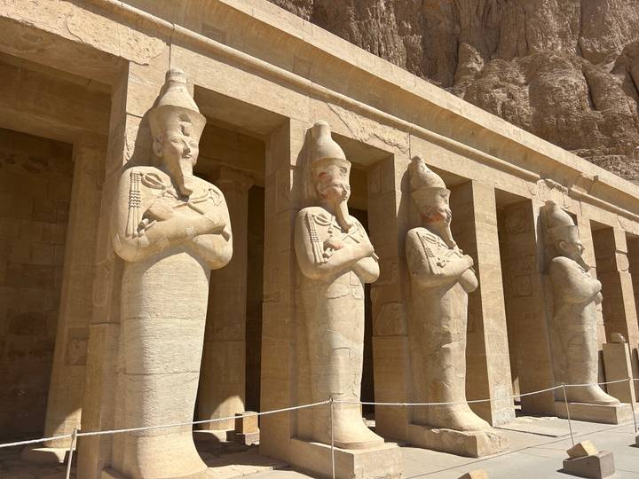 Four standing Osiride statues guard the facade of Hatshepsut's Mortuary Temple in bright sun.