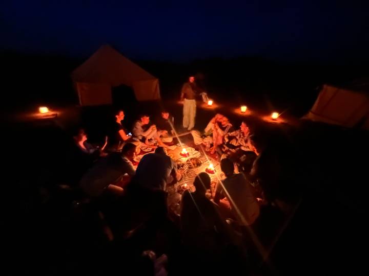 Travelers gather around a glowing campfire for dinner, surrounded by candle lanterns at a desert campsite.