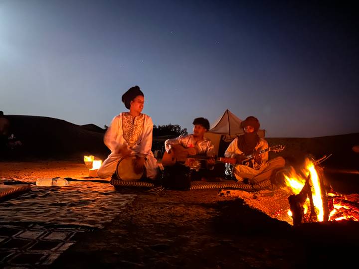 Musicians play drums and guitar beside a campfire with a night sky overhead at a desert camp.