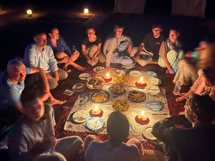 A large group sits cross-legged around platters of Moroccan food lit by candles on patterned carpets.