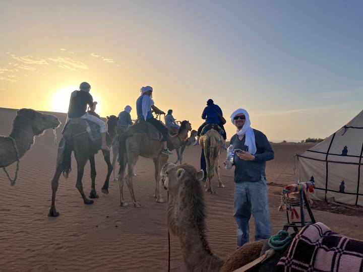 A convoy of camels and riders is silhouetted against the setting sun beside a desert camp.