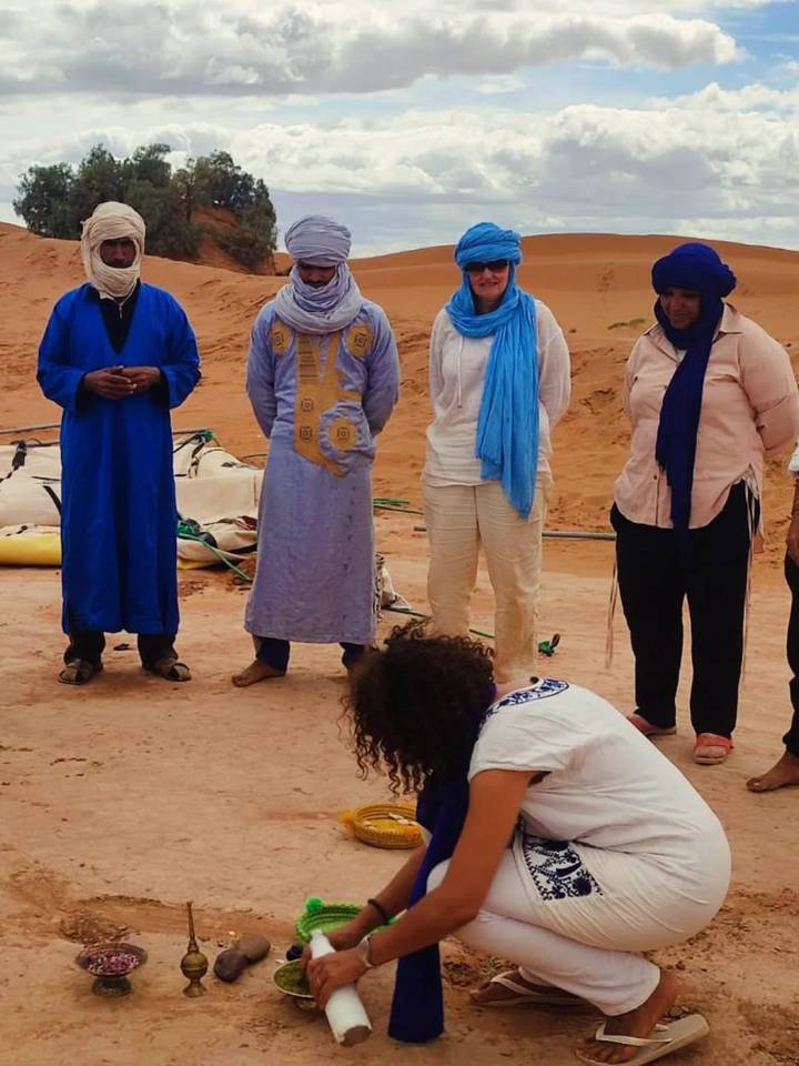 Travelers listen to a guide in traditional dress beside camel gear on desert sand.