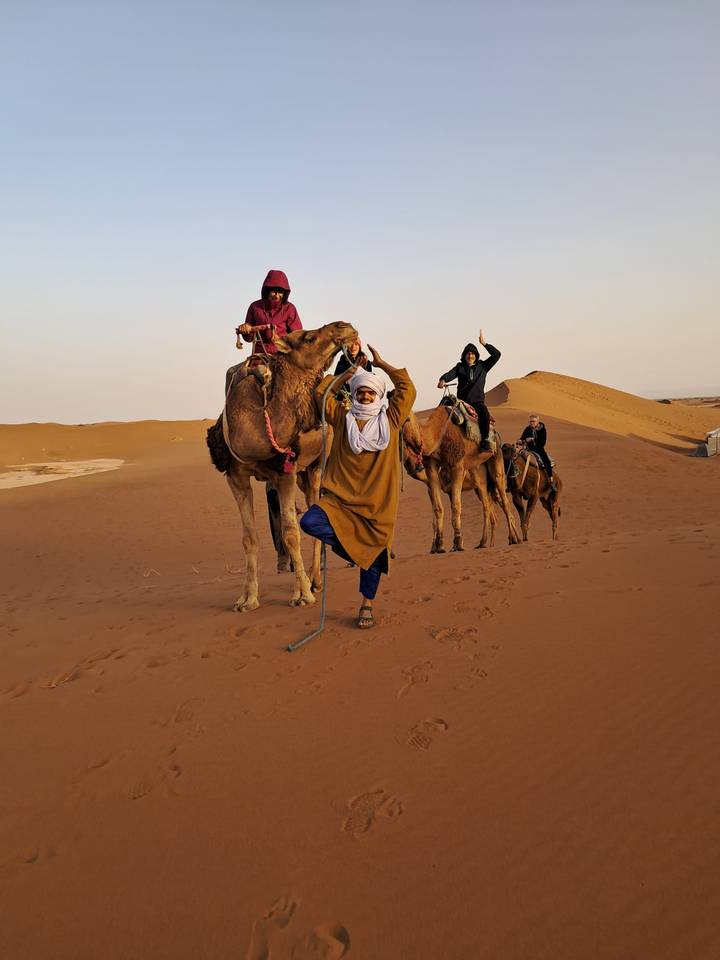 Camel riders strike playful yoga poses atop their mounts on reddish desert dunes at dawn.