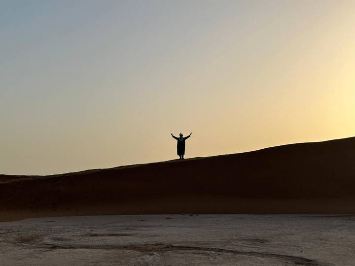 A lone silhouetted figure raises arms atop a high sand dune against a hazy desert sunset.