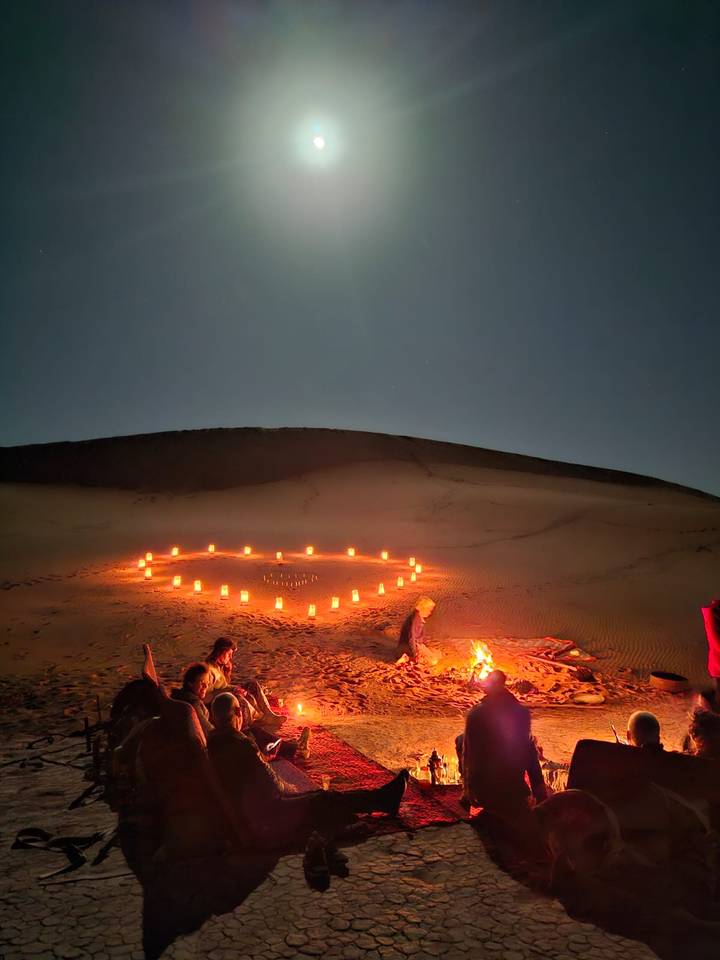 Candles arranged in a glowing heart shape light the sands while travelers sit by a campfire under the night sky.