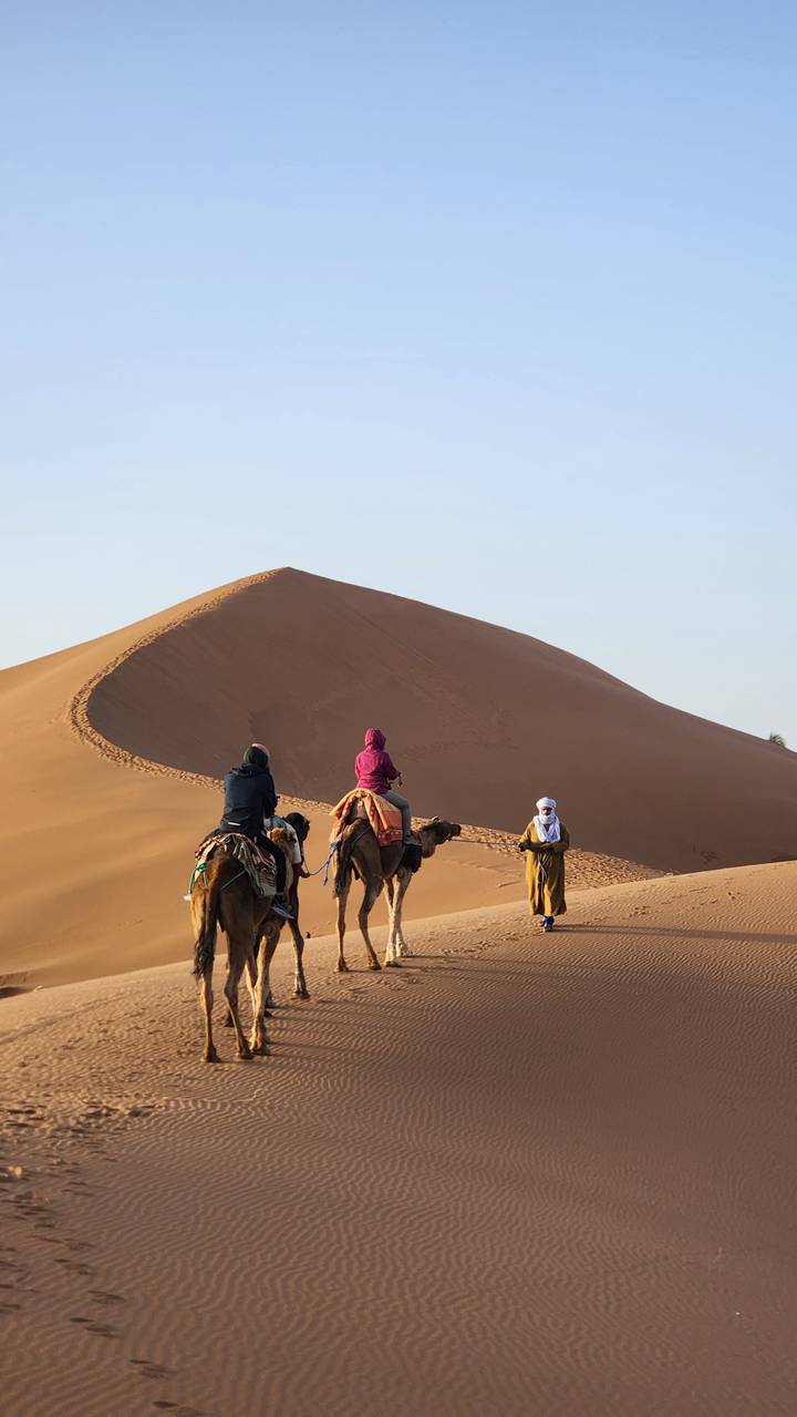Two travelers on camels approach a towering golden sand dune while a guide waits below.
