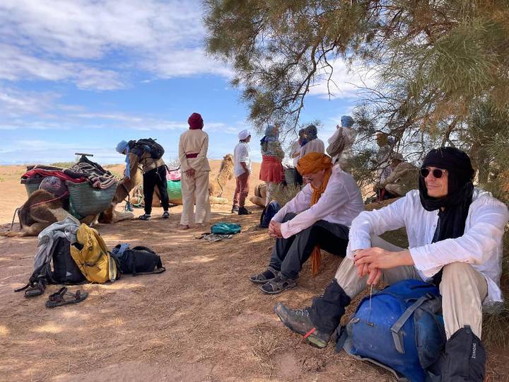 Desert trekkers resting under sparse shade with camels and gear nearby.