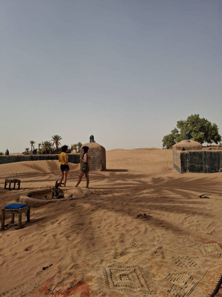 Two travellers exploring a rustic desert settlement surrounded by sand and palms.