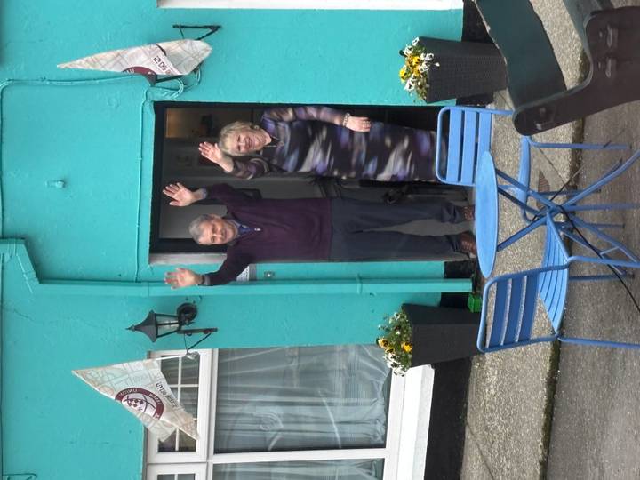 An older couple wave cheerfully from the doorway of a brightly painted turquoise house.