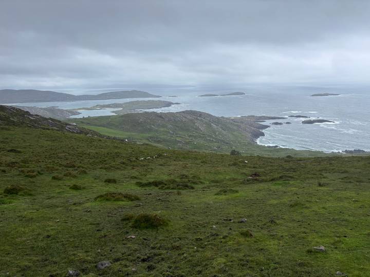 Wide view of rugged Irish coastline with rolling green hills and offshore islets under a grey sky.