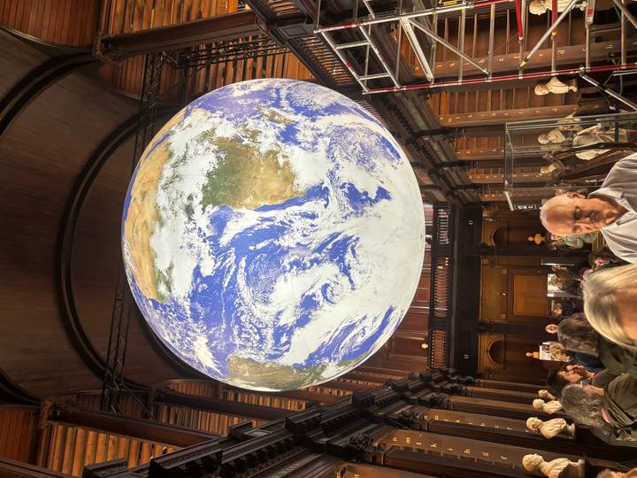 Large suspended globe art piece inside a grand wooden hall filled with visitors looking upward.