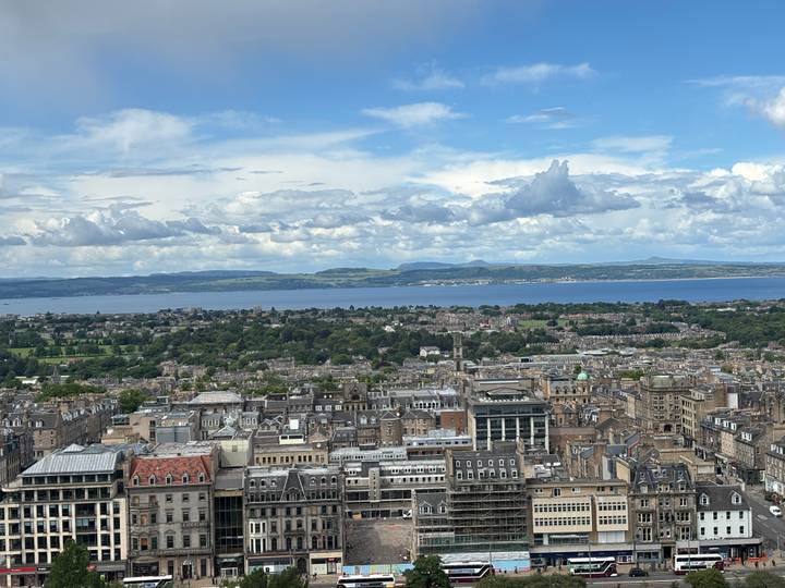 Panoramic cityscape of Edinburgh with rooftops, greenery and the Firth of Forth under patchy clouds.