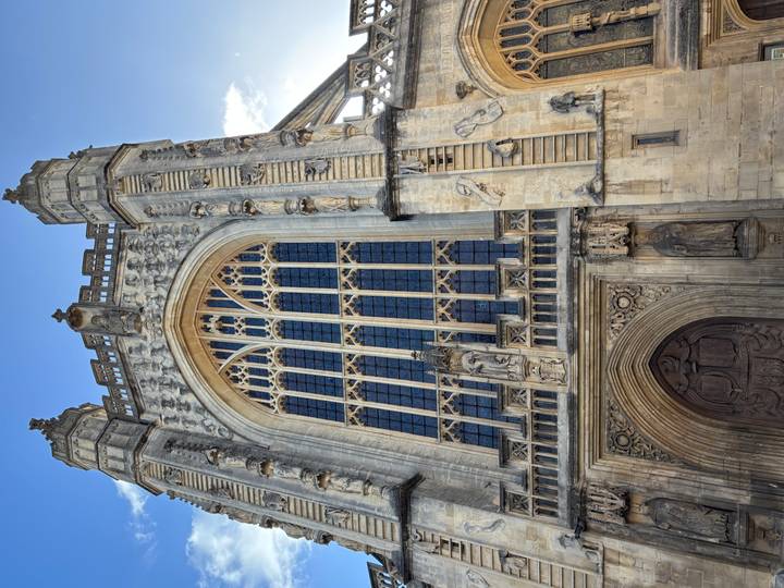 Gothic façade of Bath Abbey with towering windows, carvings and a bright blue sky background.