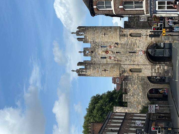 Stone medieval gatehouse with towers and heraldic shields set against a blue sky.