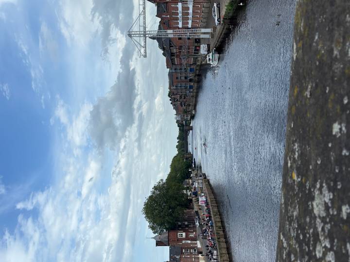 Calm river flanked by brick buildings and moored boats under a soft blue sky.