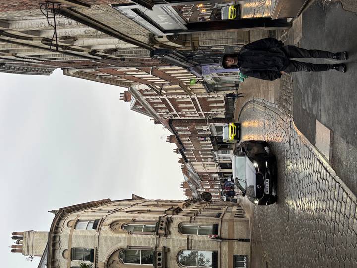 Rain-slick medieval street with a lone pedestrian in dark clothing and wet cobblestones reflecting light.