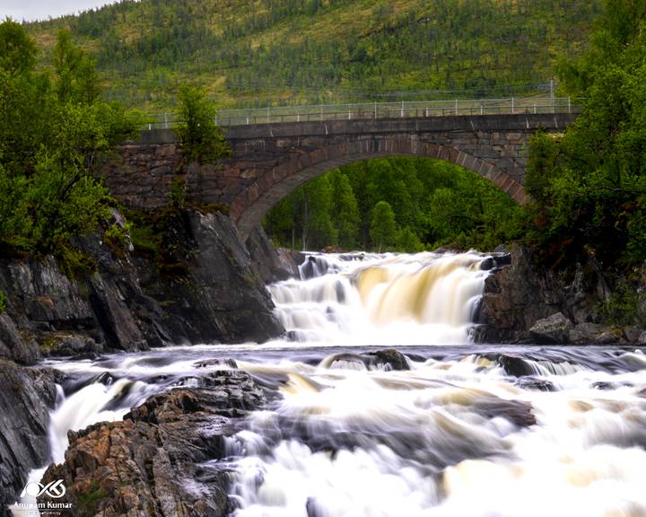 Silky waterfall plunges beneath a stone arch bridge amid lush Norwegian forest.