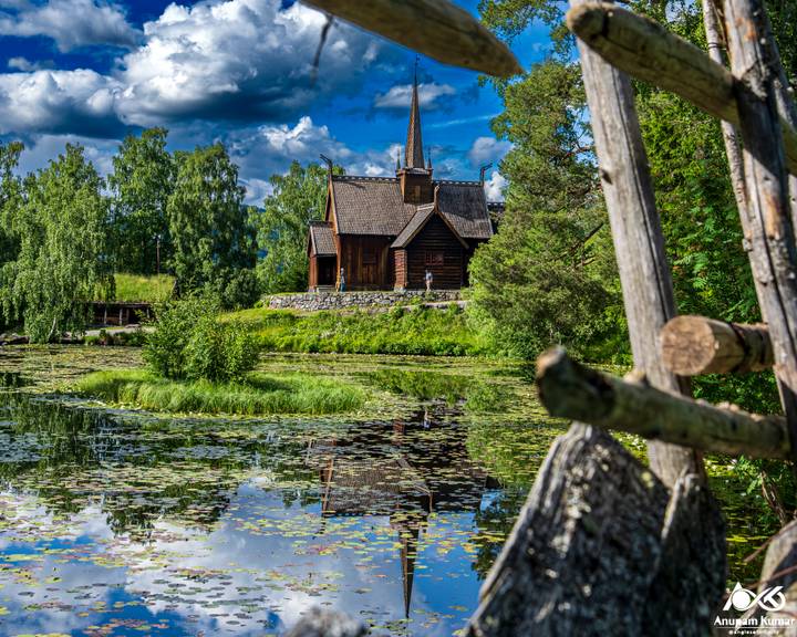 Traditional stave church reflected in a lily-covered pond surrounded by lush greenery and dramatic clouds.