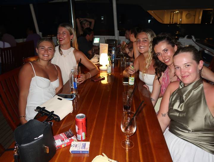 Group of friends enjoying drinks around a polished wooden table on a boat at night.