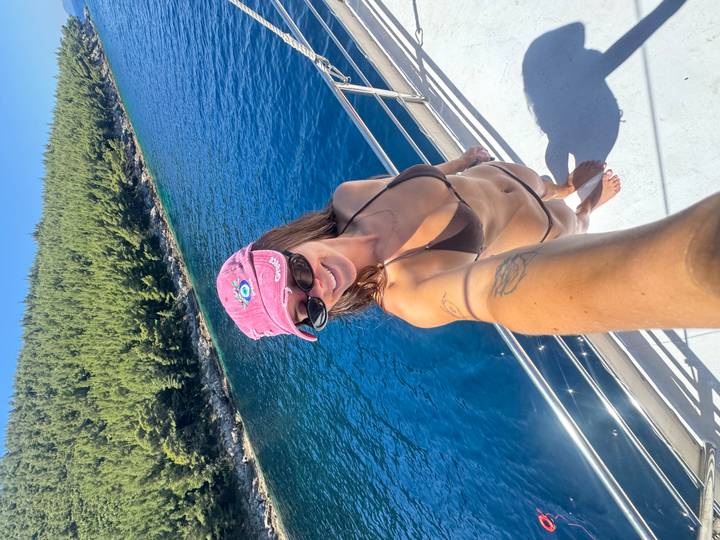 Woman in a bikini taking a selfie on a yacht deck with deep blue sea and pine-covered shoreline behind.