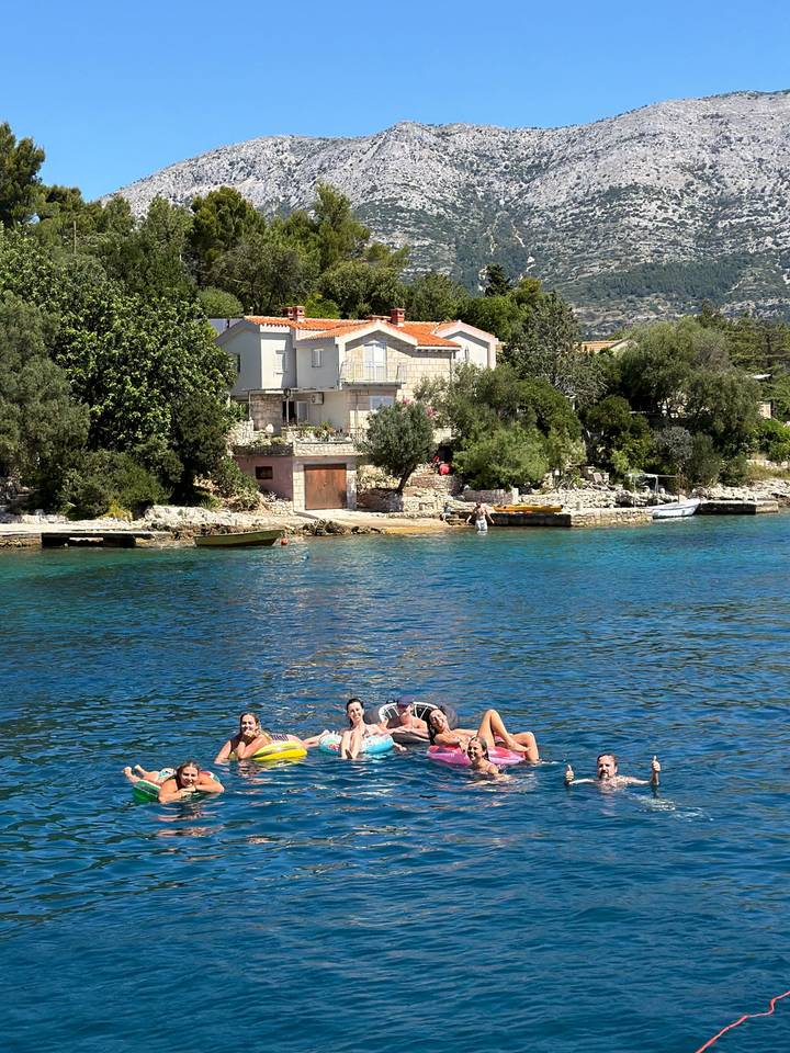 Small group floating on an inflatable raft in turquoise coastal water near stone houses.