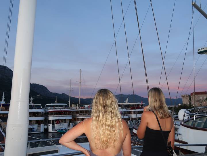 Two women admire pastel sunset skies over a harbor filled with modern cruise vessels.