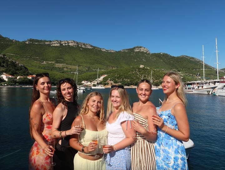 Six friends pose with drinks against a backdrop of green hills, yachts and clear blue sky.