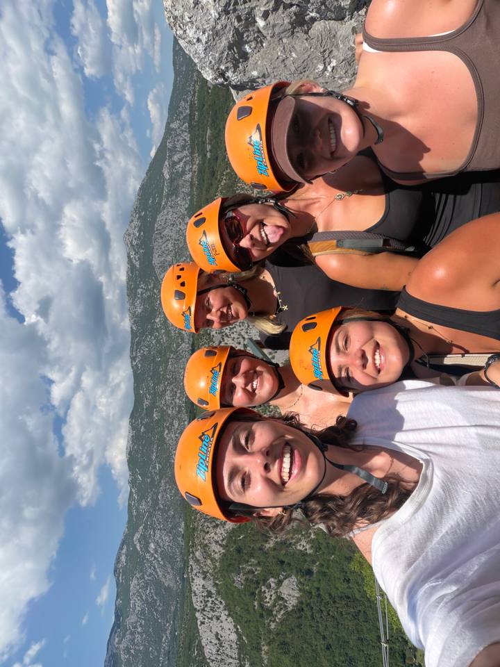 Smiling adventurers wearing orange helmets take a selfie with rugged cliffs behind.
