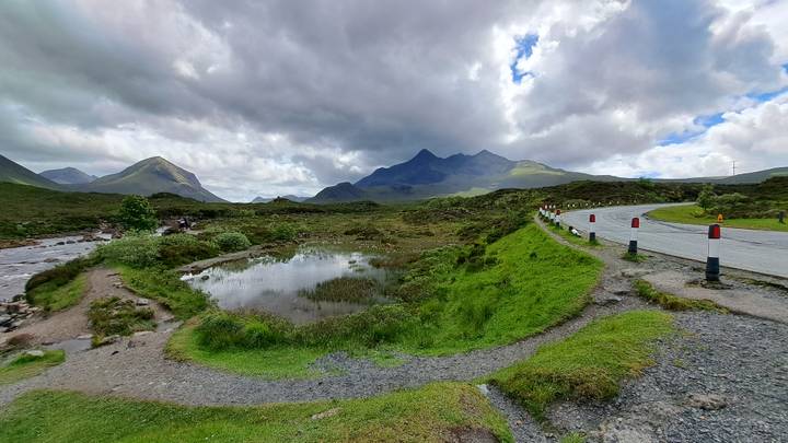 Moody Highland scene with small pond, winding road and mountains beneath heavy clouds.