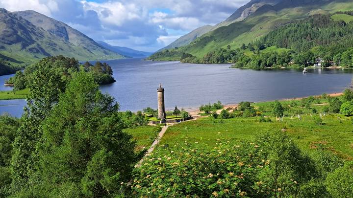 View over Glenfinnan Monument at Loch Shiel surrounded by green mountains and forest.