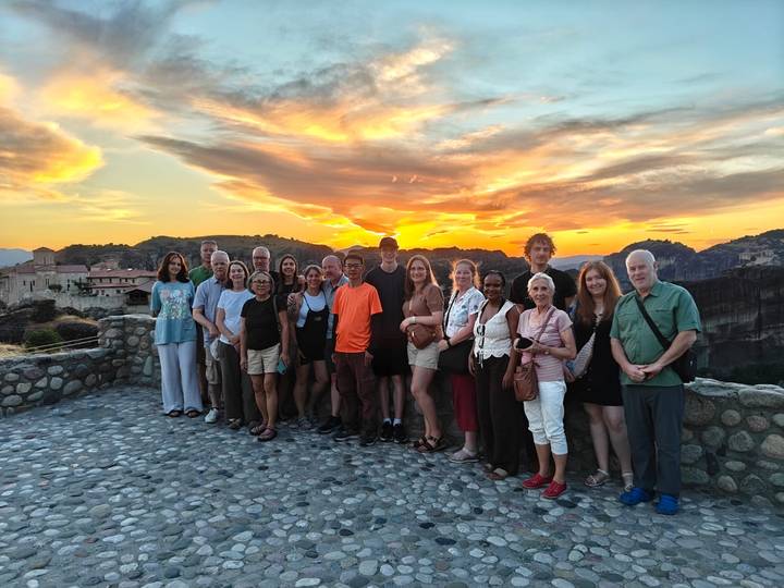 Large tour group posing together at sunset with the dramatic rock pillars of Meteora behind.