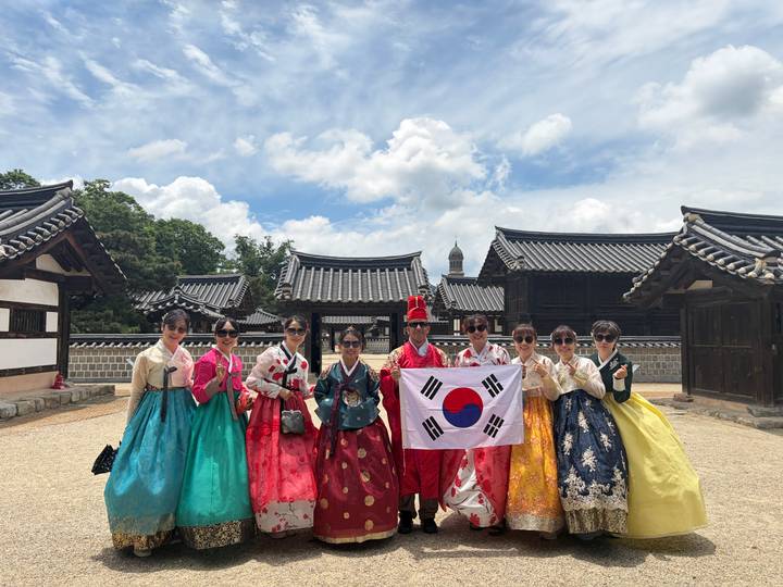 Group of visitors in vibrant hanbok posing with the Korean flag inside a traditional village courtyard.