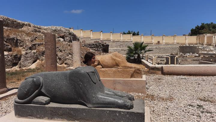 Visitor exploring an open-air archaeological site with a black granite sphinx statue and sandy ruins