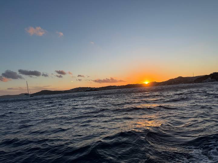 Sun setting over choppy sea waters with distant hills and a lone sailboat