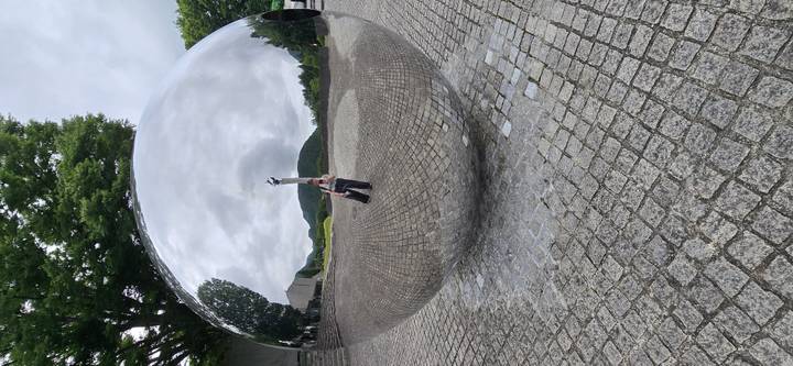 Large mirrored sphere sculpture reflecting surrounding plaza and hills with visitor visible in reflection
