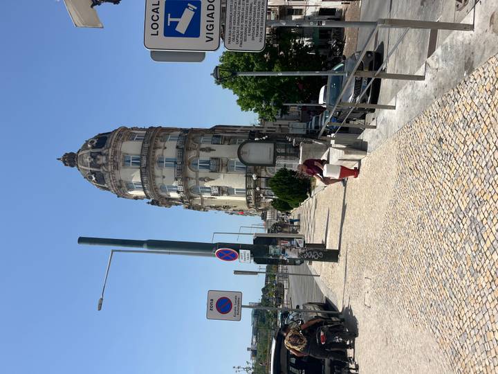 Historic turreted building rises beside a sunlit mosaic sidewalk and passing traffic