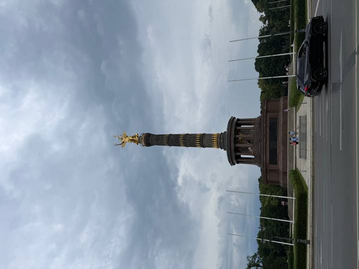 Tall Victory Column with gold statue rising against a gray sky, small visitors and traffic at the base