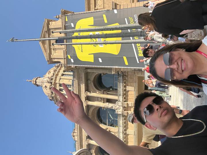 Two travellers smiling for a selfie in front of an ornate domed building and large event banner, with other visitors on the steps behind.