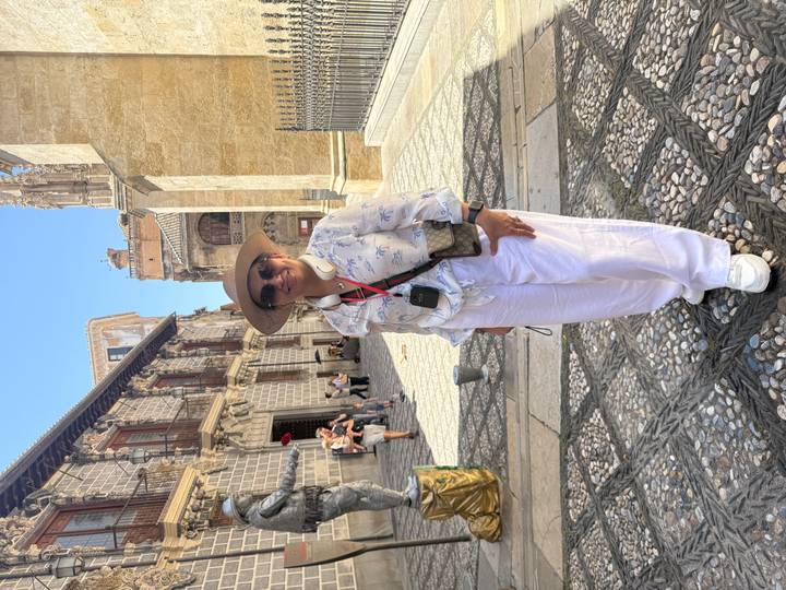 Traveller in hat and headphones posing in a historic cobblestone courtyard lined with ornate stone buildings.